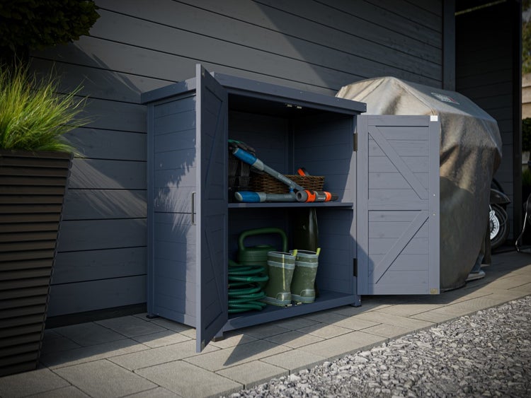 Geöffneter Gartenschrank aus Holz mit Gartengeräten wie Gießkanne und Gartenschlauch auf einer gepflasterten Fläche.