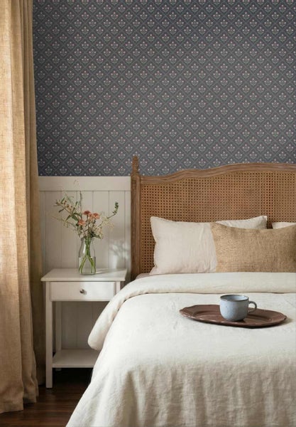 Schlafzimmer mit kleingemusterter Tapete über weißer Holzverkleidung, Bett mit Rattankopfteil, weißem Nachttisch und hellen Textilien.