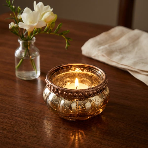 Ein brennendes Teelicht in einem runden Windlicht aus Glas auf einem Holztisch mit einer Blumenvase und einer Serviette.