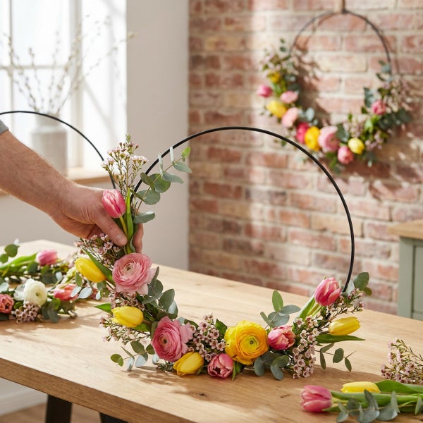 Person dekoriert einen runden Metallring mit Blumen wie Tulpen, Ranunkeln und Eukalyptuszweigen als frühlingshafte Wanddekoration.