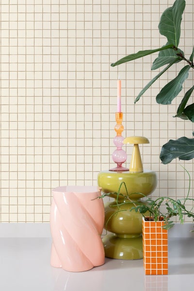 Szene mit einem rosa Hocker, einem grünen Beistelltisch, einer Kerze und einer Pflanze vor einer Wand mit quadratischem Muster.