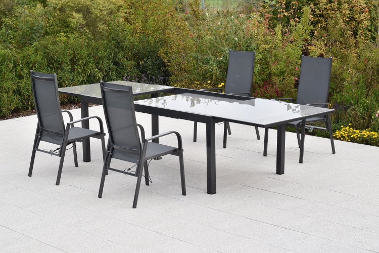 Gartensitzgruppe mit einem ausziehbaren Esstisch mit Glasplatte und vier Hochlehner-Stühlen auf einer hellen Steinterrasse im Garten.
