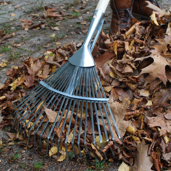 Ein metallener Laubbesen mit Holzstiel, der in einem Haufen herbstlicher Blätter steckt.