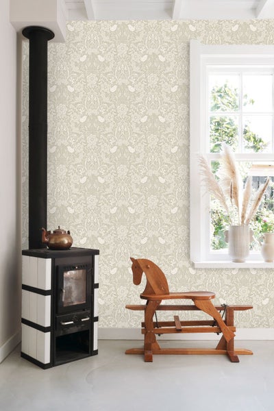 Heller Raum mit Vliestapete mit floralem Muster und Vögeln, Kaminofen, Holzschaukelpferd und Fenster mit Pampasgras.