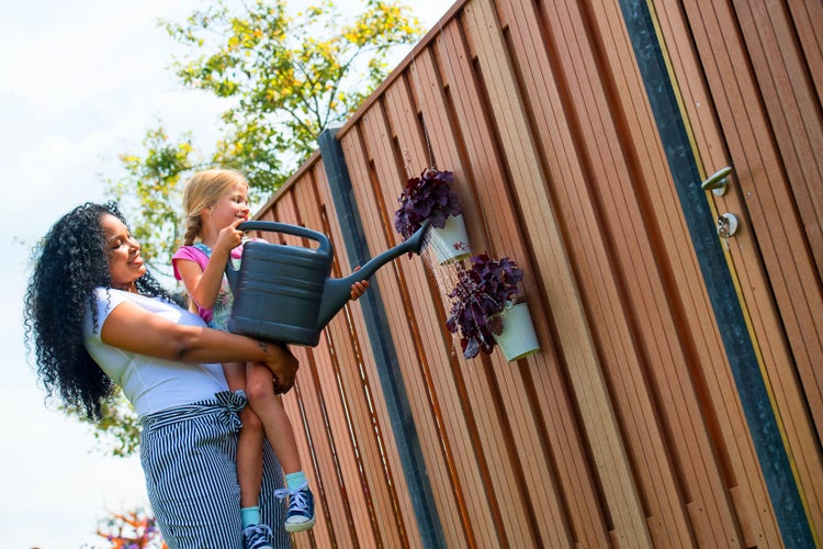 Frau und Kind gießen Pflanzen an einem Holzzaun mit einer Gießkanne.