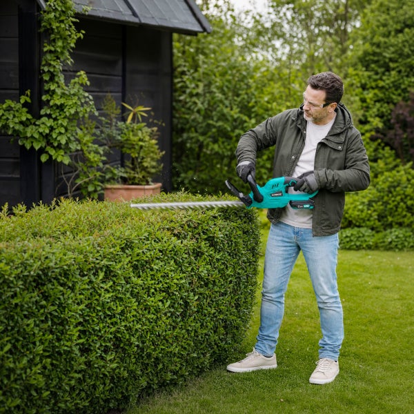 Ein Mann schneidet eine grüne Hecke mit einer türkisfarbenen Akku-Heckenschere von Vonroc in einem Garten.