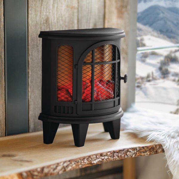 Ein elektrischer Kaminofen im Gusseisen Look mit realistischem Flammeneffekt steht auf einer Holzbank vor einem verschneiten Winterfenster.