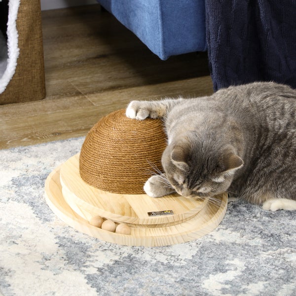 Katzenspielzeug mit Kratzkuppel aus Sisal auf rundem Holzsockel mit Kugelbahnen, eine Katze spielt damit auf einem Teppich im Wohnzimmer.
