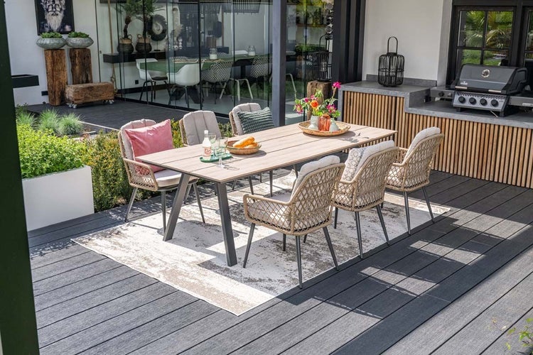 Modernes Gartenmöbel-Set auf einer Terrasse. Holztisch mit Metallgestell, sechs geflochtene Stühle mit Sitzkissen und ein Außenteppich.