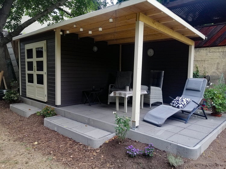 Modernes Gartenhaus aus Holz mit überdachter Terrasse, anthrazitfarbenen Wänden, Doppeltür mit Glas und Außenmöbeln auf Fliesen.