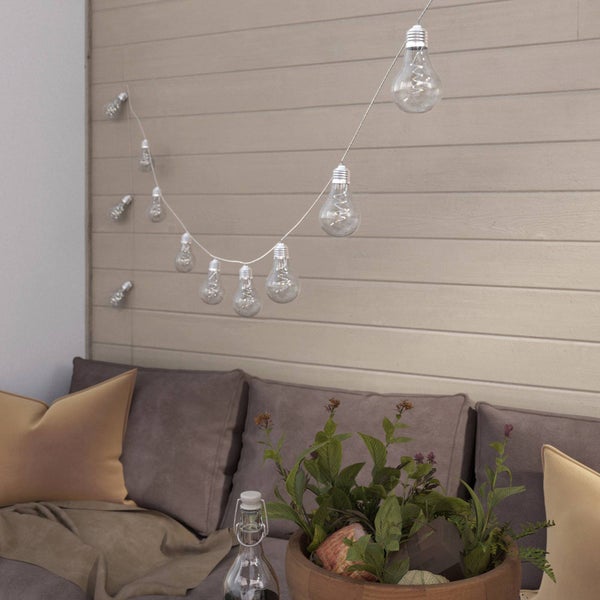 LED-Lichterkette mit klaren Glühbirnen vor einer Holzpaneelwand über einem Sitzbereich mit Kissen und Pflanzen.