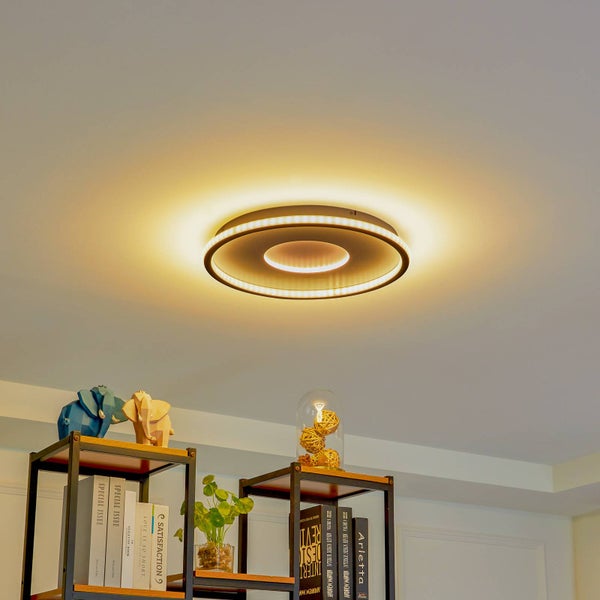 Moderne runde LED-Deckenleuchte mit zwei warmweißen Leuchtringen in einem hellen, organisierten Raum über einem Regal mit Büchern und Pflanzen.