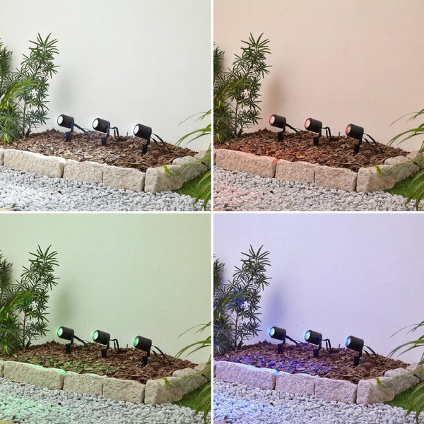 Drei LED-Gartenstrahler in einem Beet mit Rindenmulch und Steineinfassung, dargestellt in den Lichtfarben Weiß, Rot, Grün und Blau.