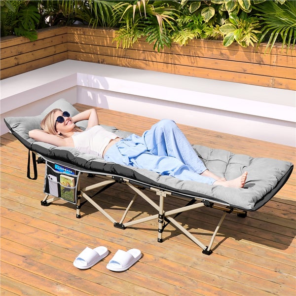 Klappbares Feldbett mit dicker grauer Matratze und Seitentasche auf einer Holzterrasse mit einer entspannten Person.