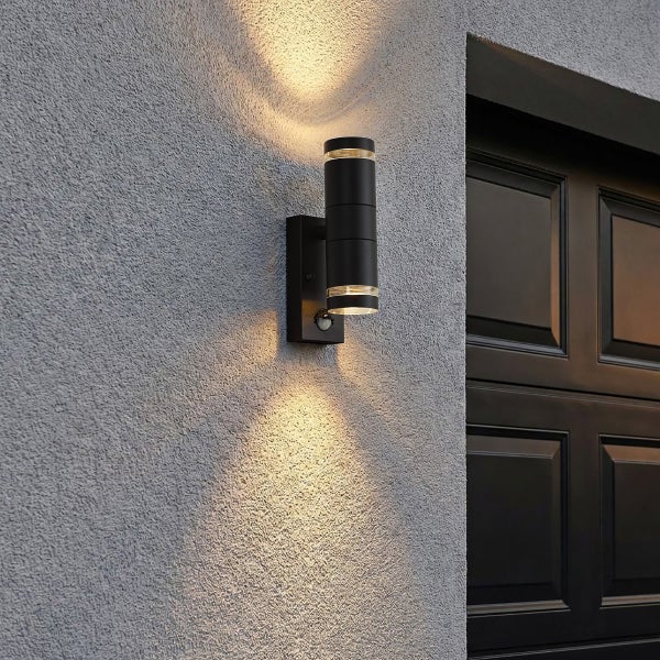 Moderne schwarze zylindrische Außenwandleuchte mit Bewegungsmelder, Up-and-Down-Lichteffekt an einer strukturierten Fassade.