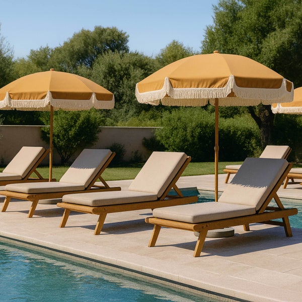 Sonnenliegen aus Holz mit beigen Auflagen und senfgelbe Sonnenschirme mit weißen Fransen an einem Pool im Garten.
