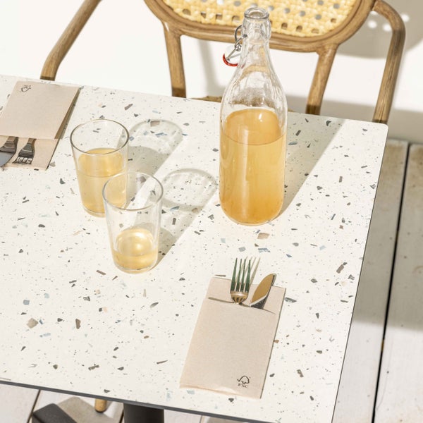 Gedeckter Tisch mit Terrazzo-Platte, Glasflasche, zwei Gläsern, Besteck in FSC-zertifizierten Papierservietten und einem Rattanstuhl.