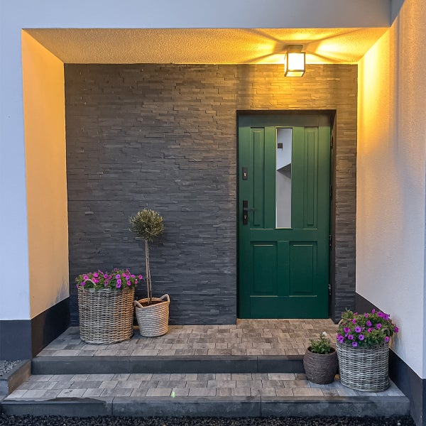 Hauseingang mit grüner Haustür, Wand mit dunklen Verblendern in Natursteinoptik, Pflanzkörben und Außenbeleuchtung.