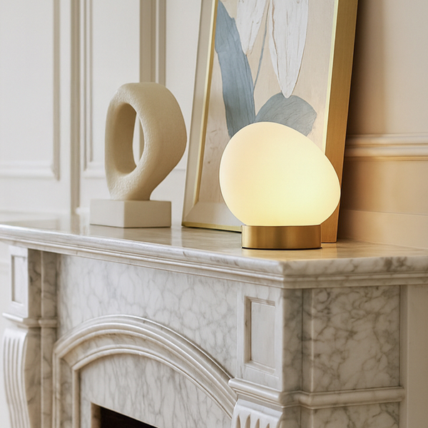 Tischleuchte in Eiform mit weißem Glas und goldfarbenem Metallfuß auf einer Kaminumrandung aus Marmor.
