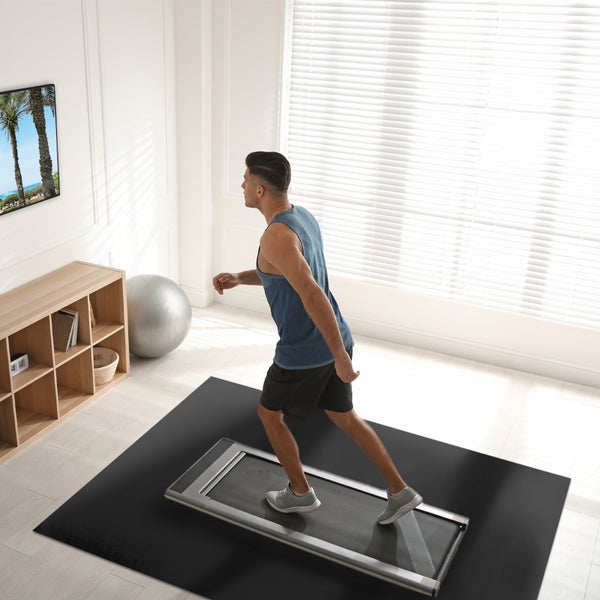 Mann trainiert auf einem flachen Laufband in einem hellen Home-Gym mit Bodenschutzmatte, Gymnastikball und Regal.