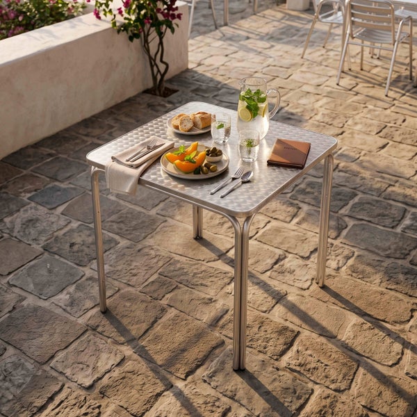 Quadratischer Bistrotisch aus Aluminium mit gemusterter Oberfläche, gedeckt mit Speisen und Getränken auf einer hellen Steinterrasse.