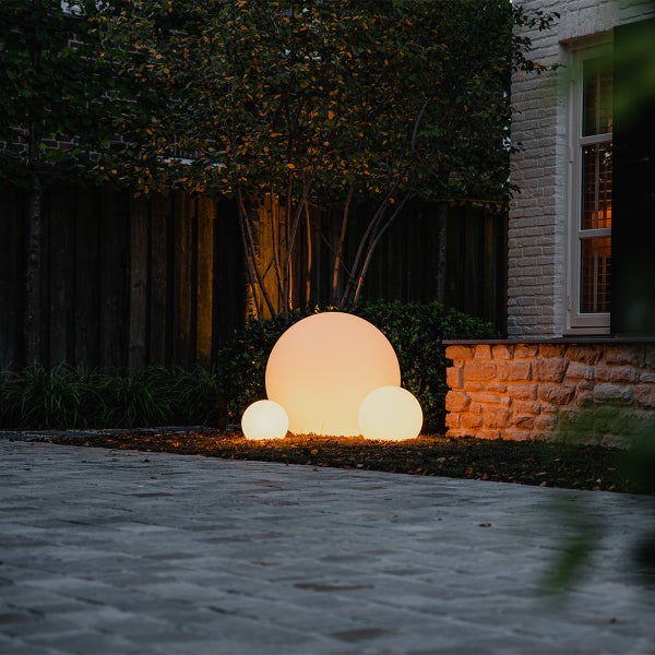 Drei beleuchtete kugelförmige Gartenleuchten in verschiedenen Größen auf Mulch vor einem Baum und einer Steinmauer bei Dämmerung.
