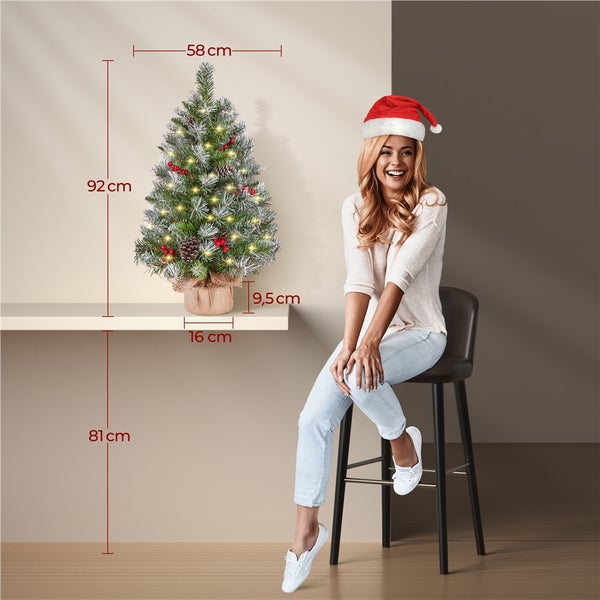 Dekorierter, beleuchteter Weihnachtsbaum mit Maßen neben einer lächelnden Frau mit Weihnachtsmannmütze