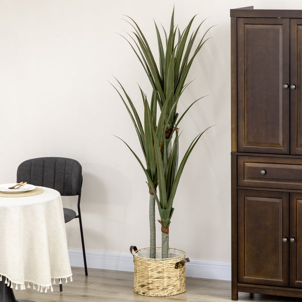 Künstliche Pflanze mit spitzen grünen Blättern in einem geflochtenen Korb, neben einem dunklen Holzschrank und einem Esstisch in hellem Raum.