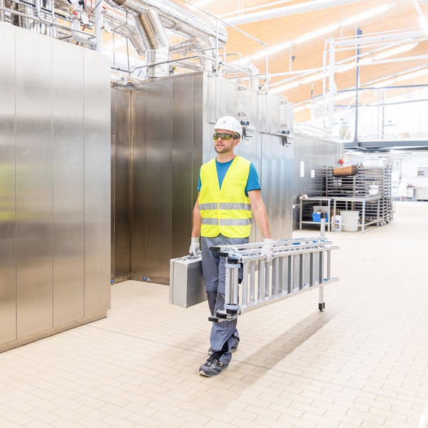 Mann in Arbeitskleidung trägt eine Leiter und einen Werkzeugkasten in einem Industriebereich.