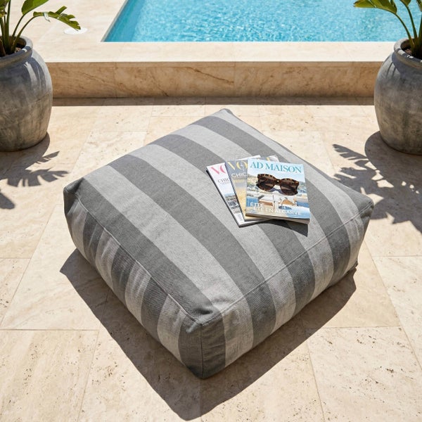 Gestreiftes Bodenkissen für den Außenbereich auf einer hellen Steinterrasse an einem Pool mit Zeitschriften und Sonnenbrille.