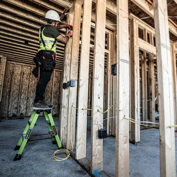 Handwerker steht auf einer Leiter und verlegt elektrische Leitungen in einem Holzständerwerk während des Hausbaus.