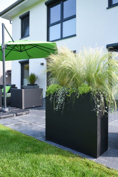 Schwarzer rechteckiger Pflanzkübel auf einer modernen hellen Terrasse mit Ziergras und Hängepflanzen neben Loungemöbeln und einem Sonnenschirm.