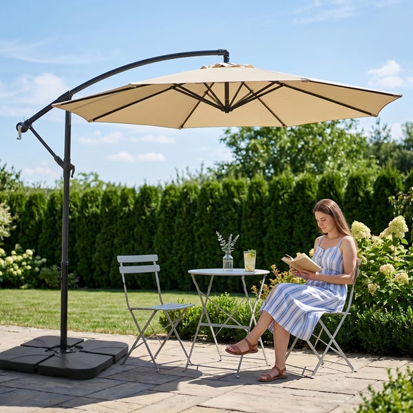 Beiger Ampelschirm über einem runden Metalltisch und Klappstühlen auf einer Terrasse, eine Frau liest ein Buch im hellen, organisierten Garten.