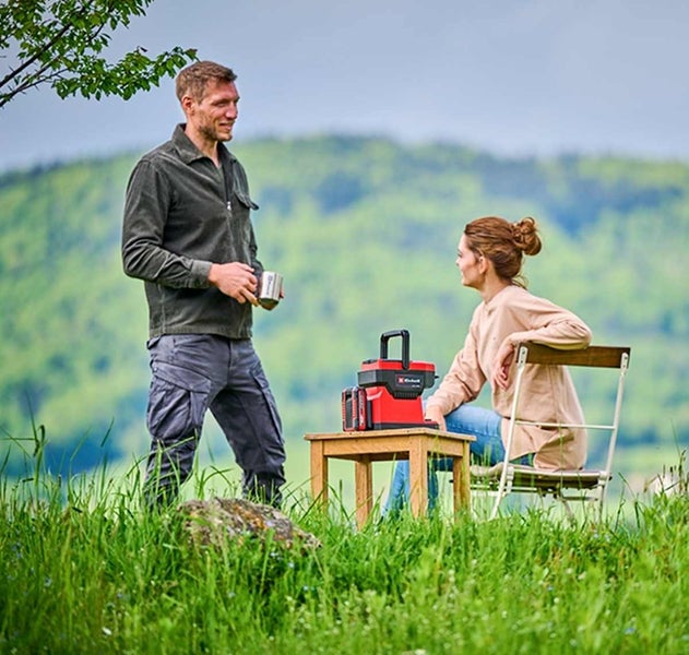 Einhell Akku-Kaffeemaschine auf einem Holztisch im Freien, eine sitzende Frau und ein stehender Mann mit Becher auf einer Wiese.