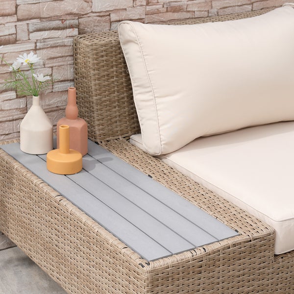 Loungesofa aus Geflecht mit hellen Polstern und integriertem Beistelltisch mit grauer Lattenoberfläche vor einer Steinwand.