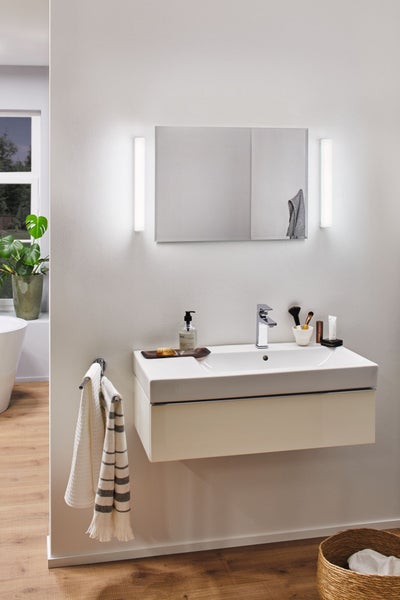 Helles Badezimmer mit wandhängendem Waschtischunterschrank, weißem Waschbecken, verchromter Armatur und Spiegel mit zwei vertikalen Lichtleisten.