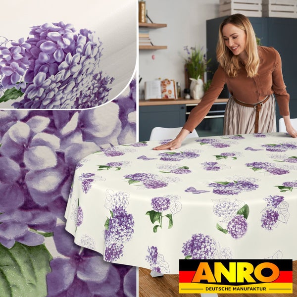 Runde Tischdecke mit lila Hortensien-Muster in einer hellen Küche. Eine Frau breitet die Decke auf einem Tisch aus. ANRO DEUTSCHE MANUFAKTUR Logo.