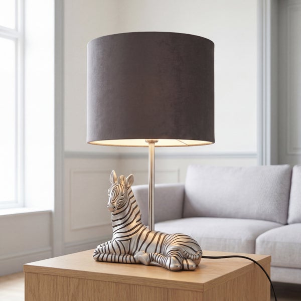 Tischleuchte mit Lampenfuß in Zebraform und zylindrischem Lampenschirm auf einem Holztisch in einem Wohnzimmer.