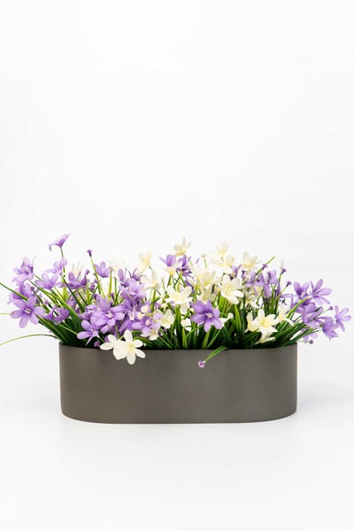 Künstliches Blumenarrangement im ovalen grauen Pflanzgefäß mit violetten und weißen Blüten.