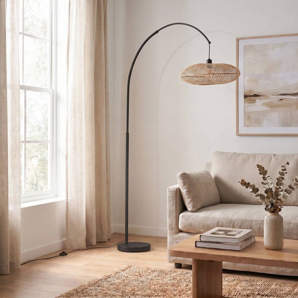 Schwarze Bogenlampe mit rundem Rattanschirm in einem hellen Wohnzimmer neben einem Sofa und einem Holzcouchtisch.
