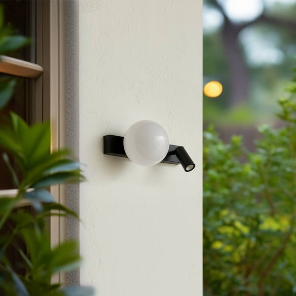 Moderne schwarze Außenwandleuchte mit weißem Kugelglas und verstellbarem Spot an einer hellen Wand, umgeben von grünen Gartenpflanzen.