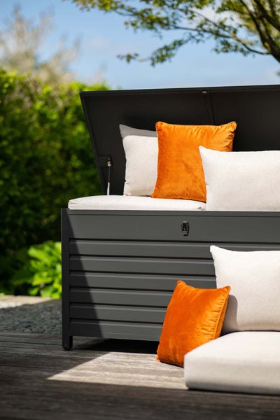 Garten-Auflagenbox aus Metall in Dunkelgrau mit geöffnetem Deckel, dekoriert mit hellen und orangefarbenen Kissen auf einer Holzterrasse.