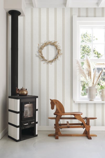 Wohnzimmer mit gestreifter Tapete, Kaminofen, Schaukelpferd aus Holz und Fensterbank mit Trockenblumen in einer Vase.