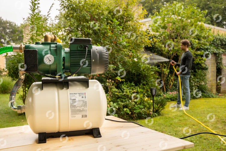 Garten mit Hauswasserwerk und Person, die mit einem gelben Gartenschlauch gießt