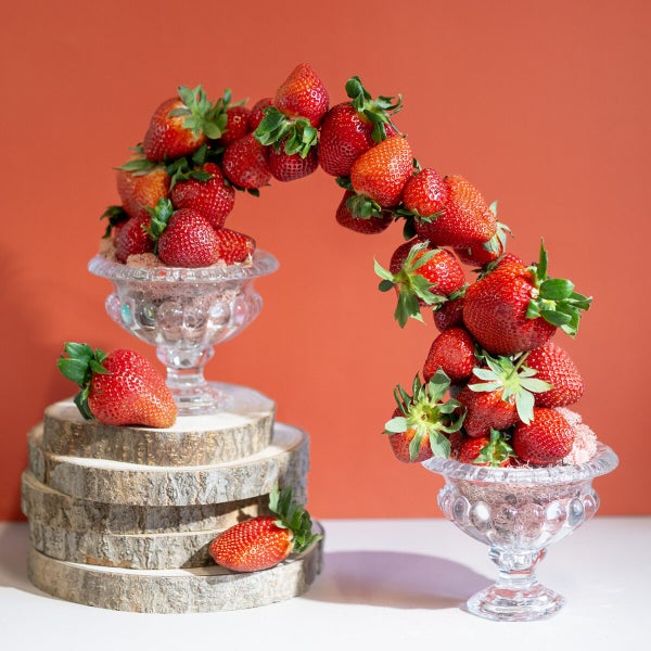Ein Bogen aus frischen Erdbeeren, der zwei mit Erdbeeren gefüllte Glasschalen verbindet, platziert auf runden Holzscheiben.