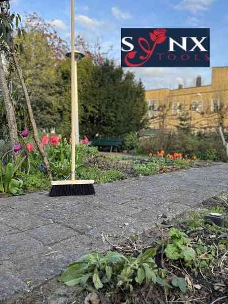 Kehrbesen mit Holzstiel auf Pflasterweg im Garten. SNX Tools Logo.