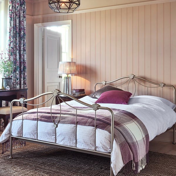 Ein Schlafzimmer mit einem Metallbett, gestreiften Wandpaneelen, Tischleuchte, Blumenvorhängen und einer gemütlichen, klassischen Einrichtung.