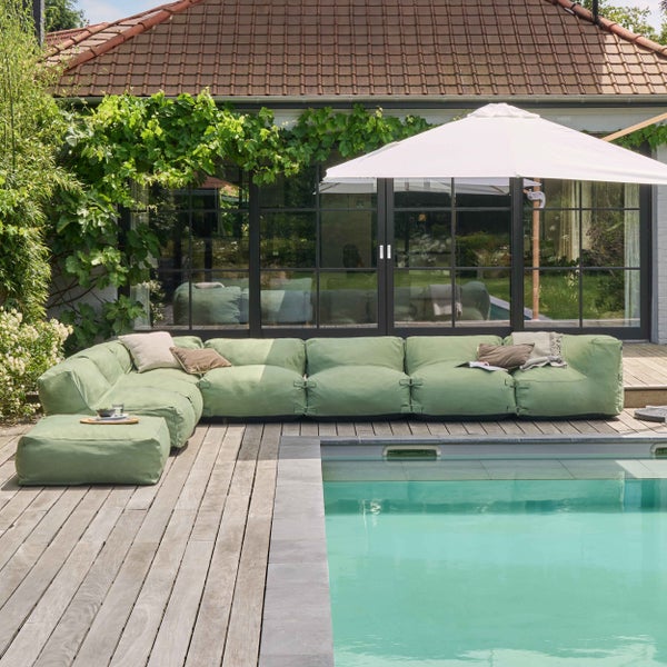 Grünes modulares Outdoor-Sofa mit Hocker auf einer Holzterrasse am Pool unter einem weißen Sonnenschirm vor einem Wohnhaus.