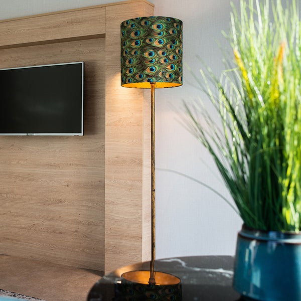 Stehleuchte mit Pfauenfeder-Muster auf dem Lampenschirm in einem Wohnzimmer neben einem Fernseher und einer Pflanze.