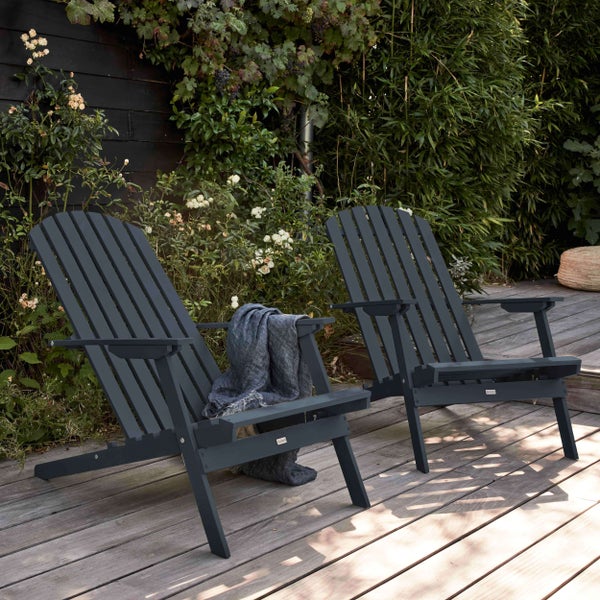Zwei dunkelgraue Adirondack-Stühle mit Lattenrücken auf einer Holzterrasse, umgeben von Gartenpflanzen und einem dunklen Zaun.